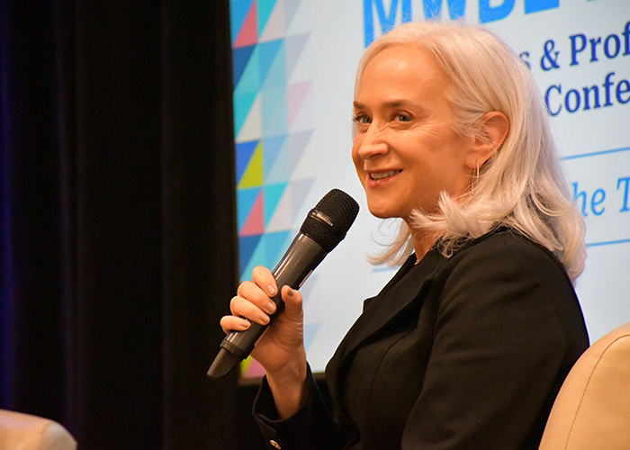 Cathy Marcus sits on stage at the MWBE Conference and speaks into a microphone.